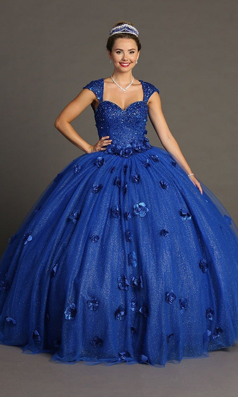 A woman wearing the Quinceanera Dress CHF3068 By Chicas in royal, adorned with 3D floral embellishments, paired with a tiara and necklace, stands against a plain gray background.